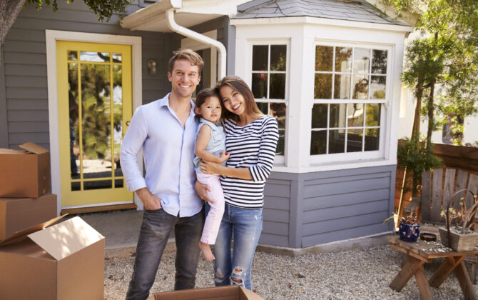 Couple with their baby standing in front of their new home with moving and relocation boxes beside them.