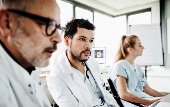 A medical student with a white coat sitting in a classroom with a teacher beside him.