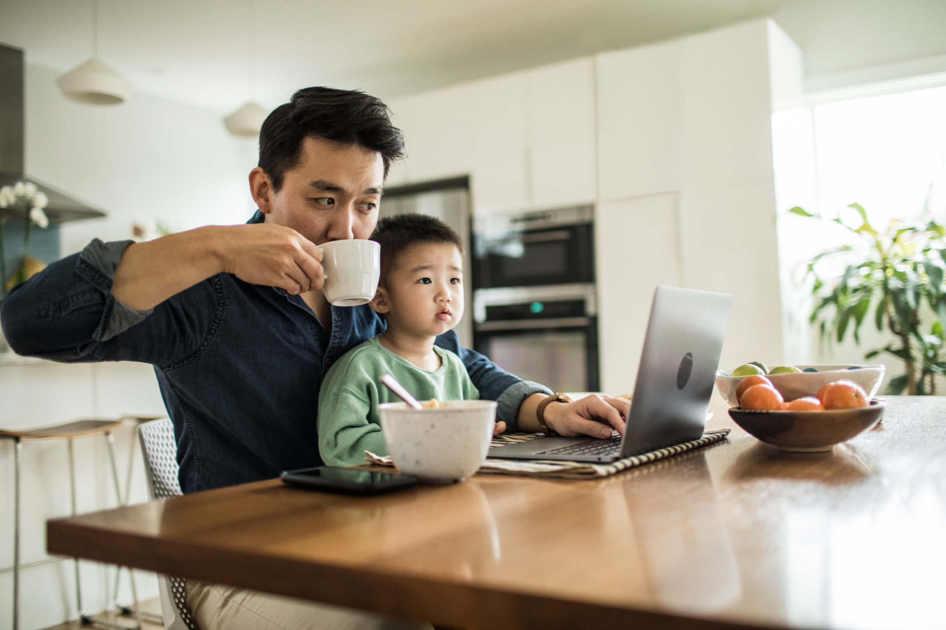 Father multi-tasking with young son at kitchen table