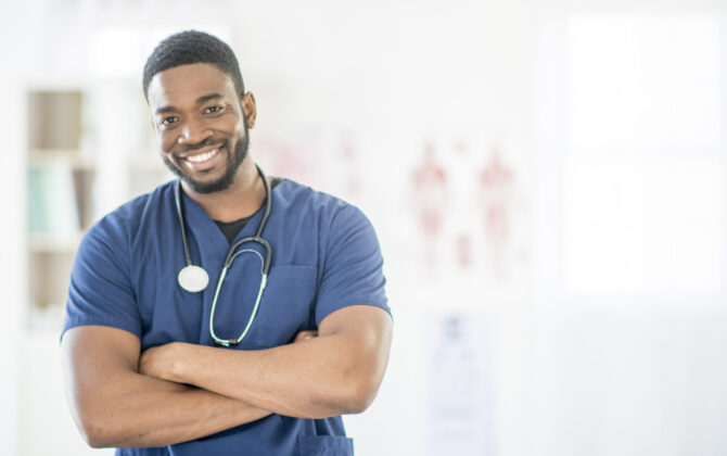 A male doctor with a blue coat and stethoscope over a shoulder, smiling wide.