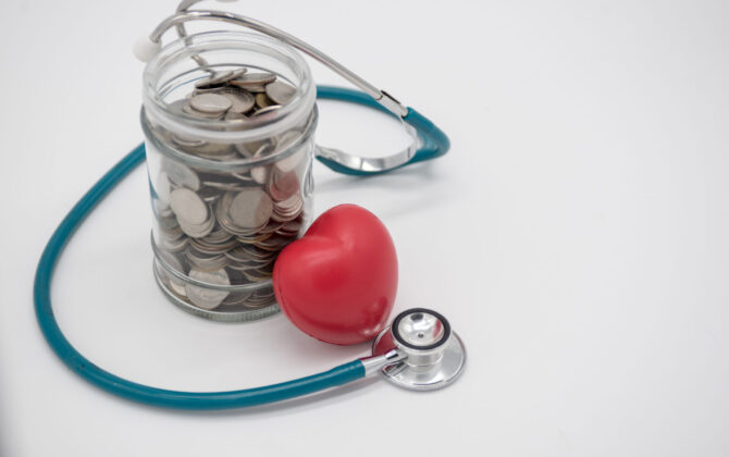 A jar filled with coins enclosed by stethoscope and a small red heart-shaped object placed near a jar.