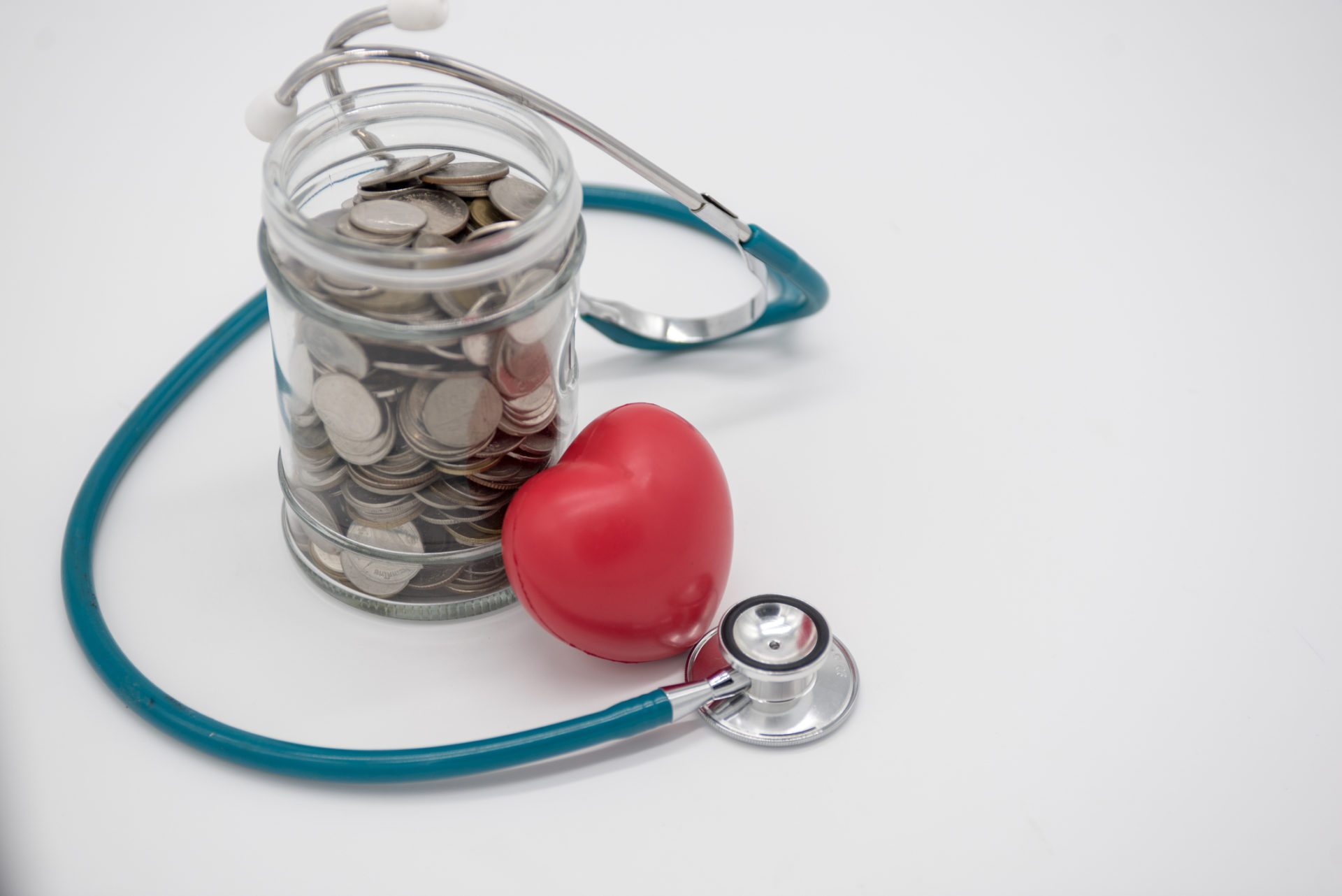 A jar filled with coins enclosed by stethoscope and a small red heart-shaped object placed near a jar.
