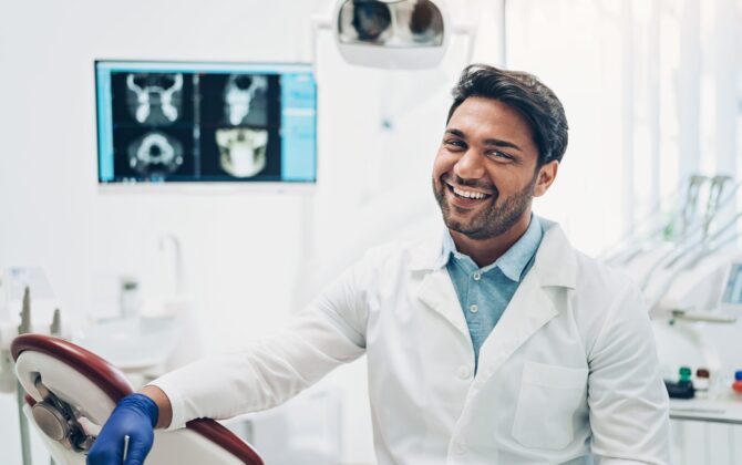 a male dentist with white coat and blue gloves, a smile on his face