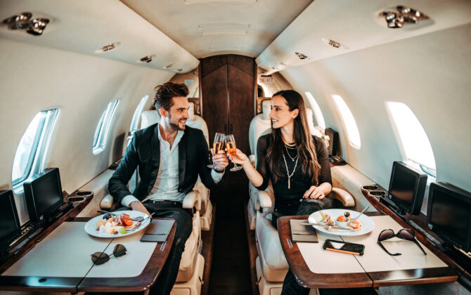 Rich couple making a toast with champagne glasses while eating canapes aboard a private jet.