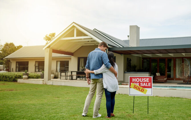 Shot of a couple standing next to a real estate sold sign at their new house