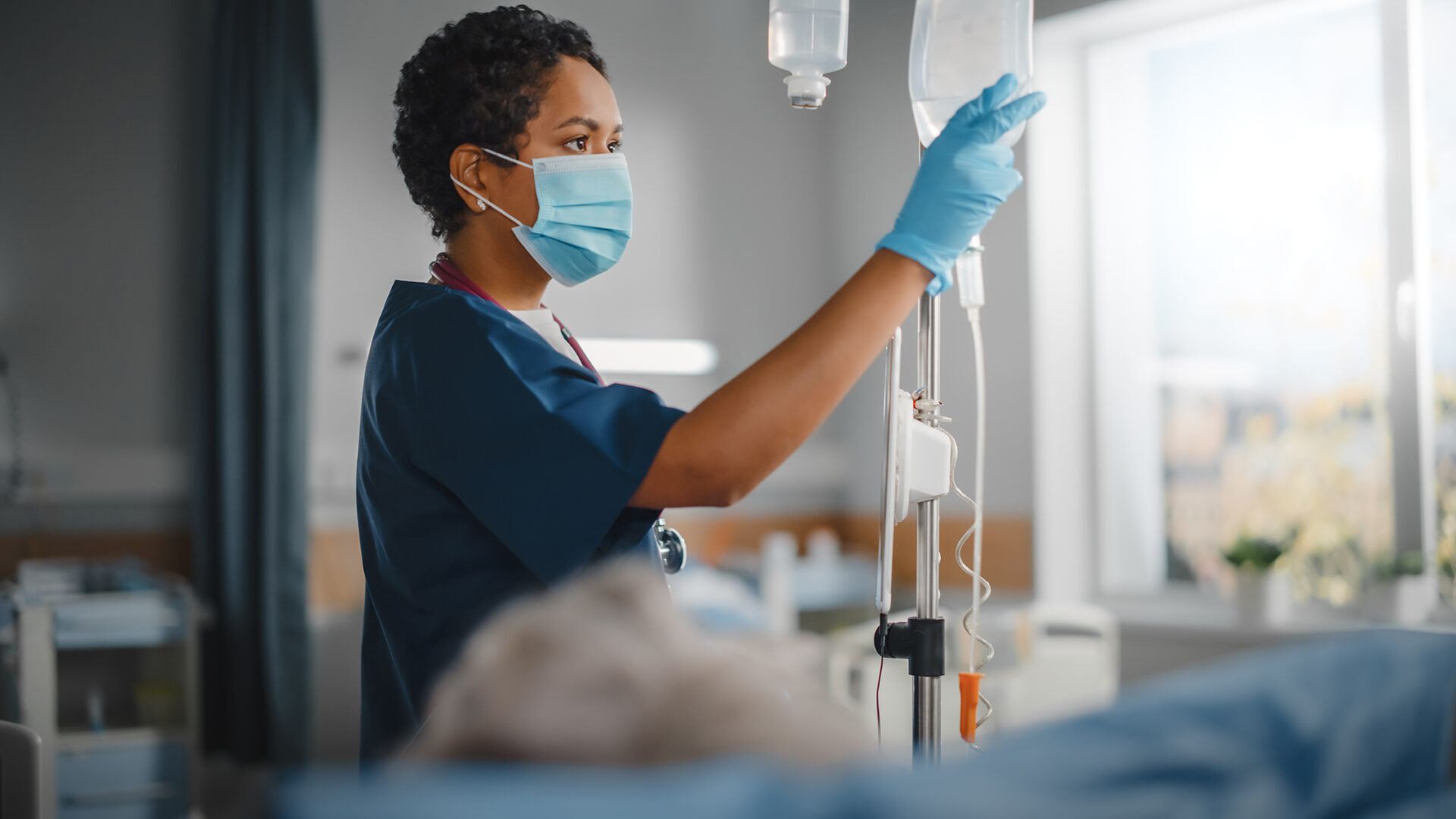 Hospital Ward: Professional Black Head Nurse Wearing Face Mask Does Checkup of Patient's Vitals, Checking Heart Rate Computer, Iv Fluids Drip Bag.