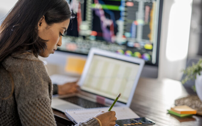 Middle Eastern woman tracking and trading stocks using laptop and desktop computer.