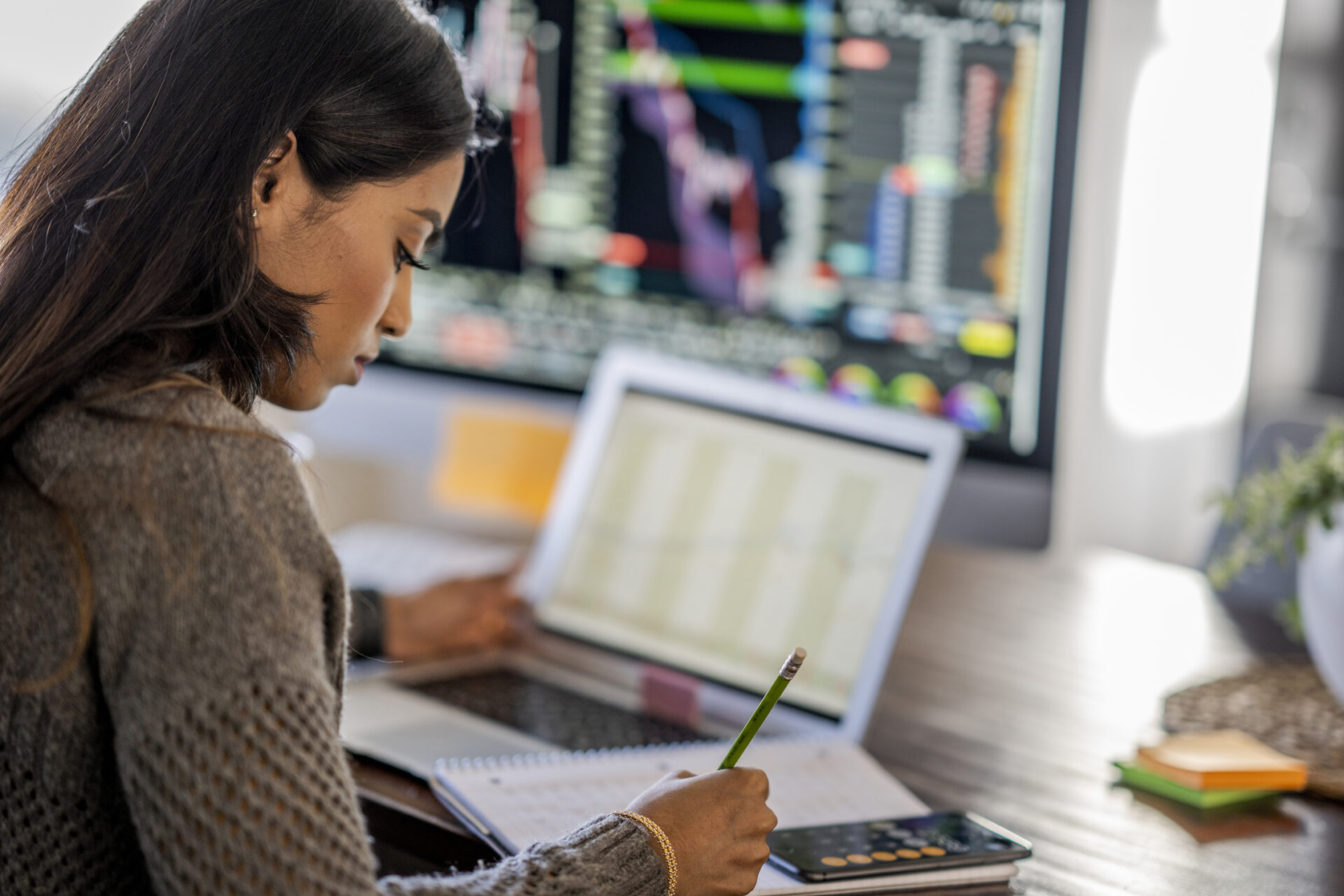 Middle Eastern woman tracking and trading stocks using laptop and desktop computer.