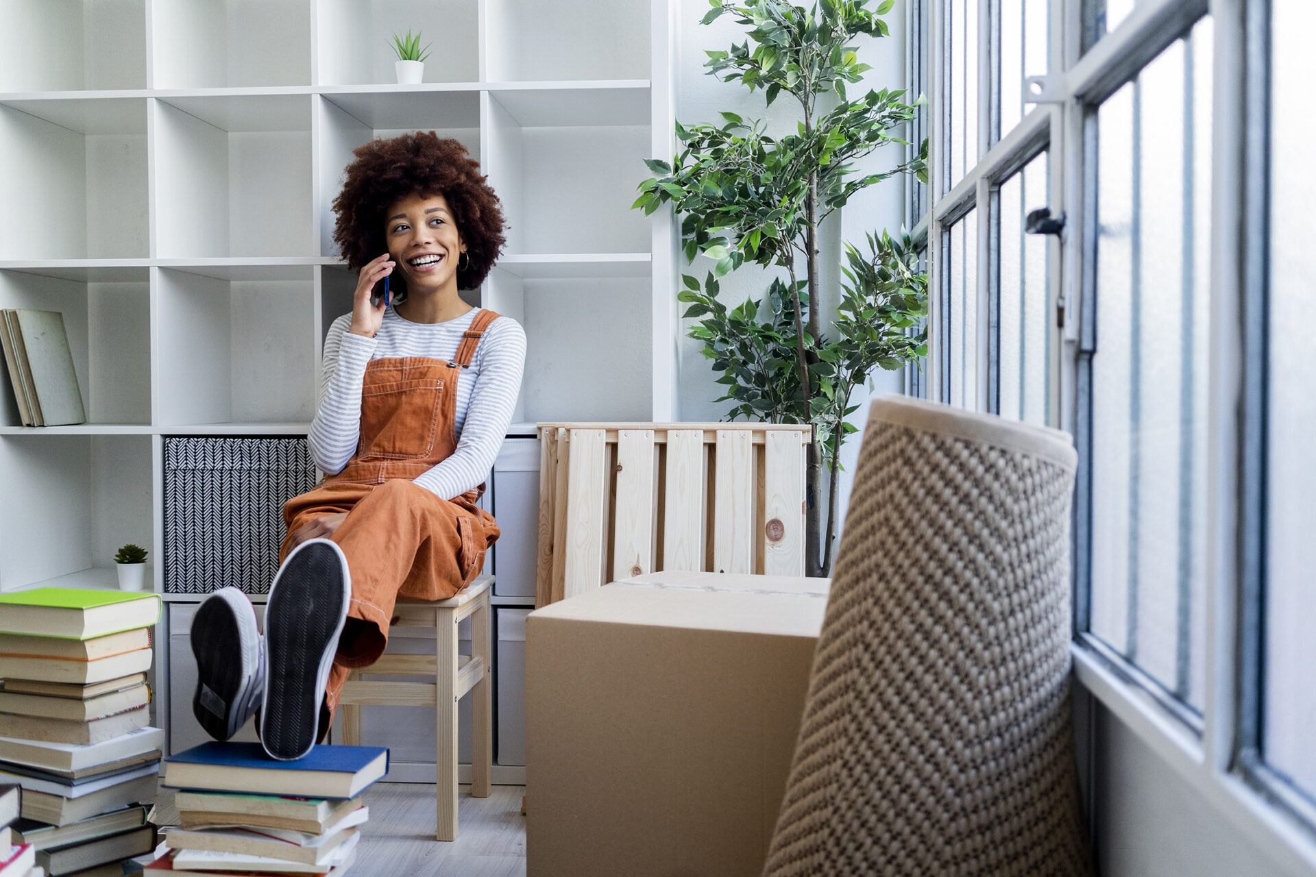 Afro woman talking on smart phone while sitting against bookshelf in new apartment
