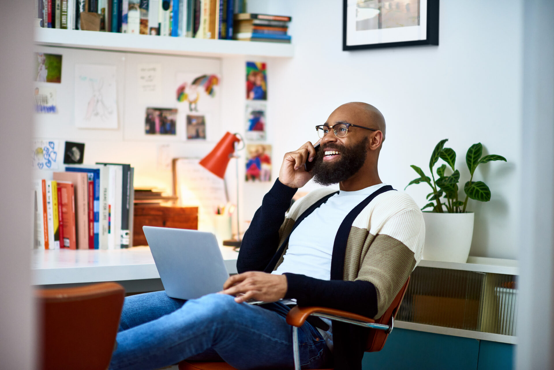 Man in his 30s sitting on chair with laptop, on phone call.