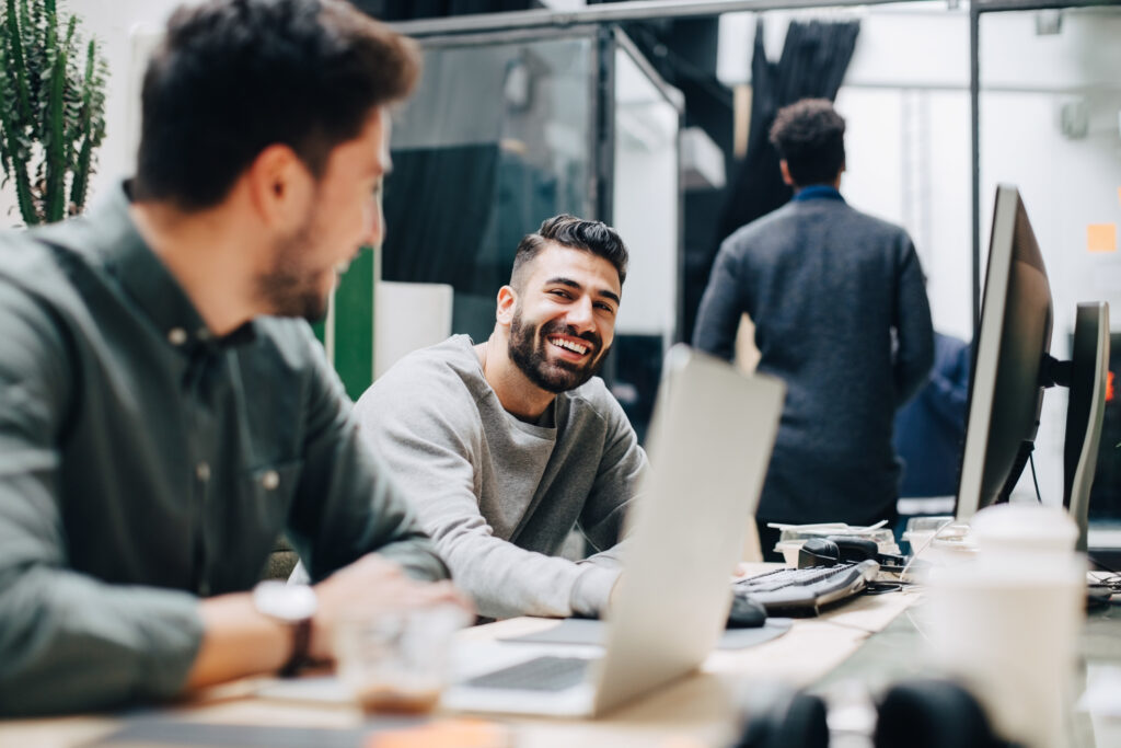 Smiling male colleagues looking at each other while sitting by desk in office