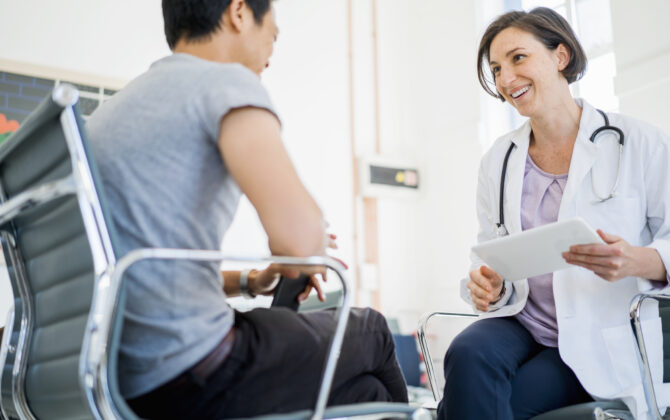 A photo of happy doctor holding digital tablet while looking at patient. Female medical professional is communicating with man. They are sitting on chairs in clinic.