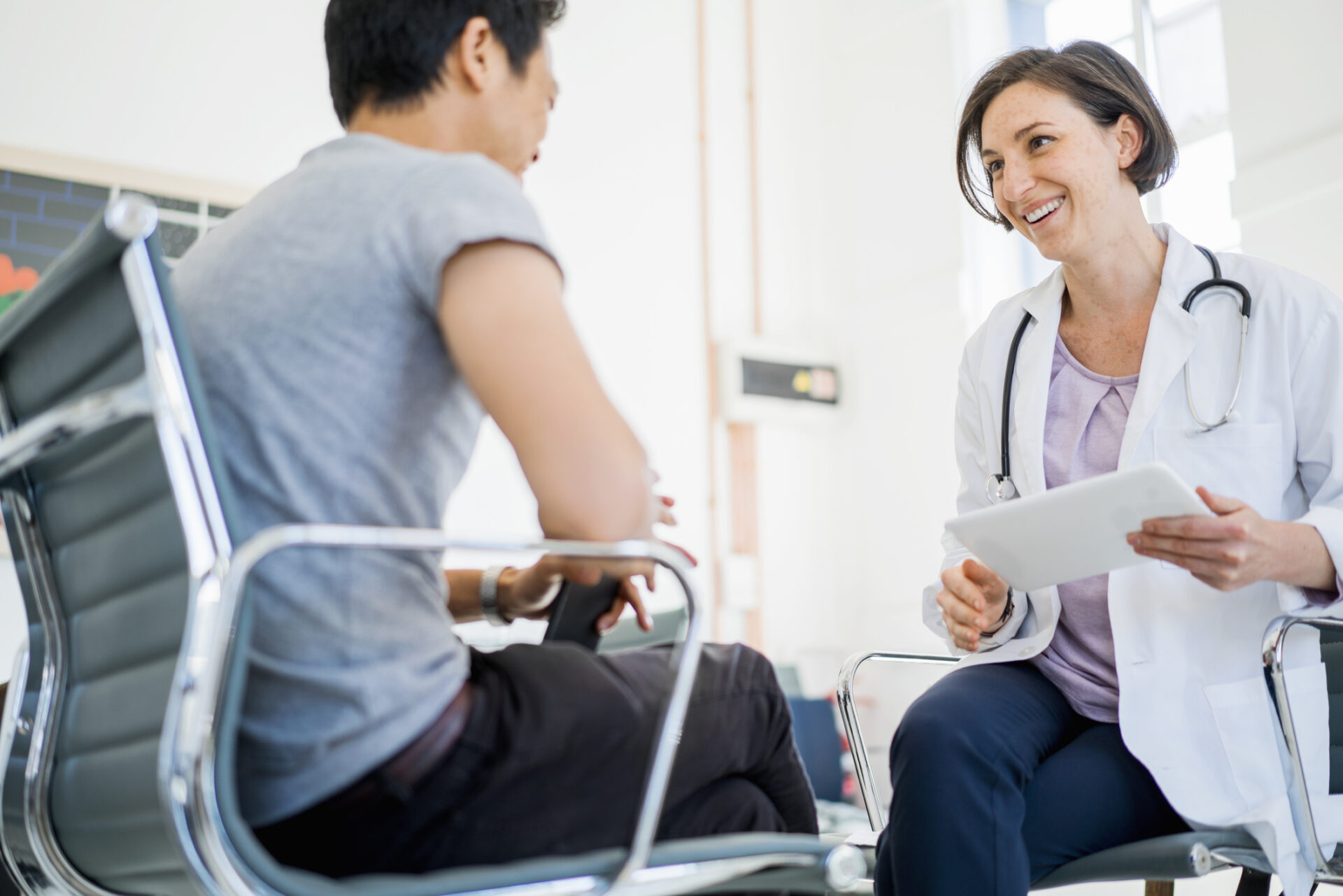 A photo of happy doctor holding digital tablet while looking at patient. Female medical professional is communicating with man. They are sitting on chairs in clinic.